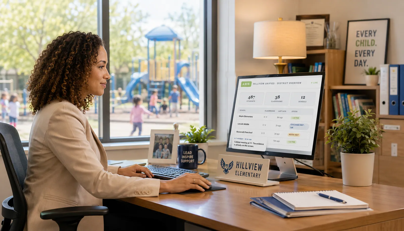 A district special education director at her desk in a school office, the ARIS District Overview dashboard open on her monitor, a playground visible through the window behind her