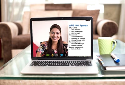 A laptop on a desk showing the ARIS 101 webinar agenda with a smiling presenter on screen